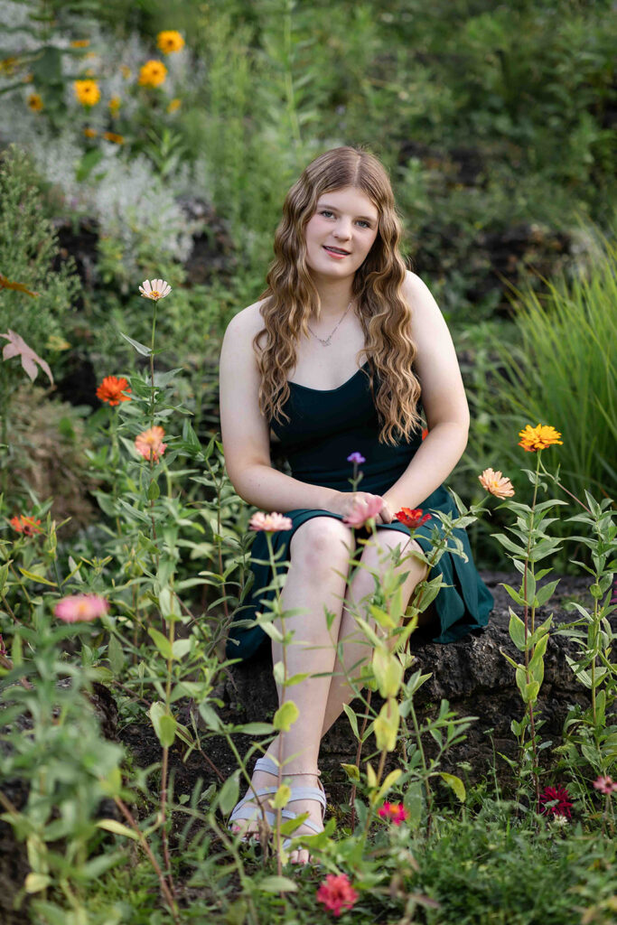 Teen girl with long wavy hair in a dark green dress sitting among flowers, leaning forward softly and smiling during birthday portrait session at Rockway Gardens in Kitchener-Waterloo.