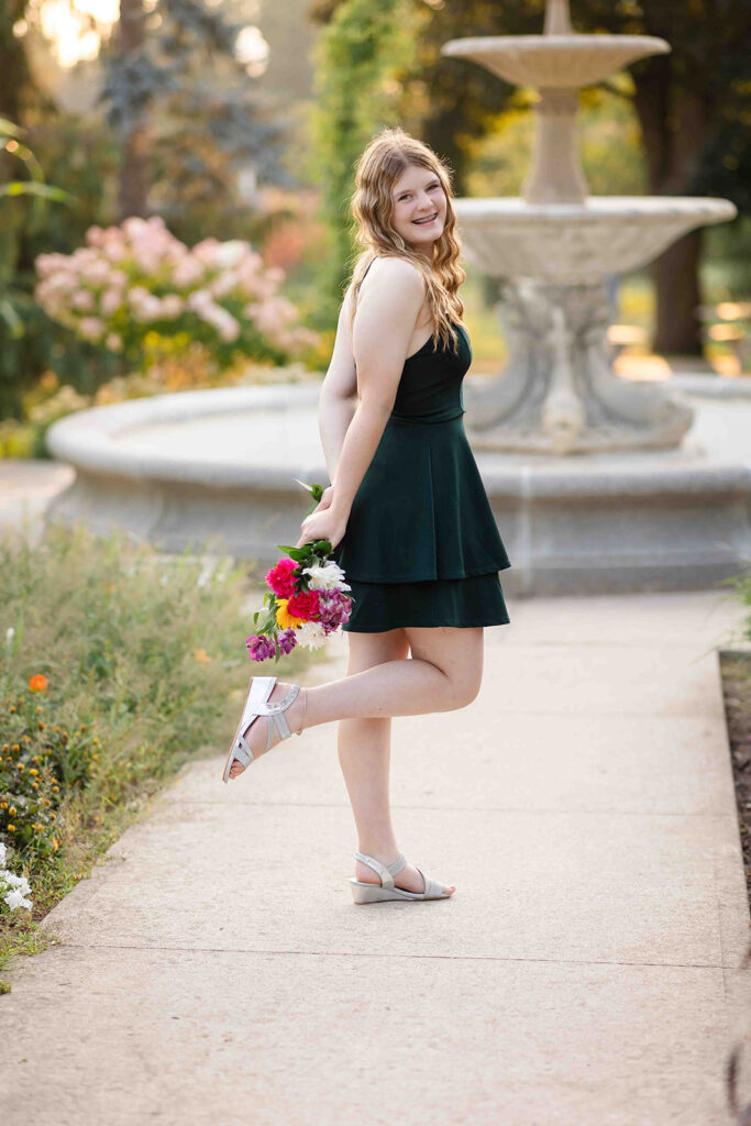 Teen girl in green dress holding a flower bouquet, looking over her shoulder with a soft smile at an outdoor portrait location in Kitchener-Waterloo.