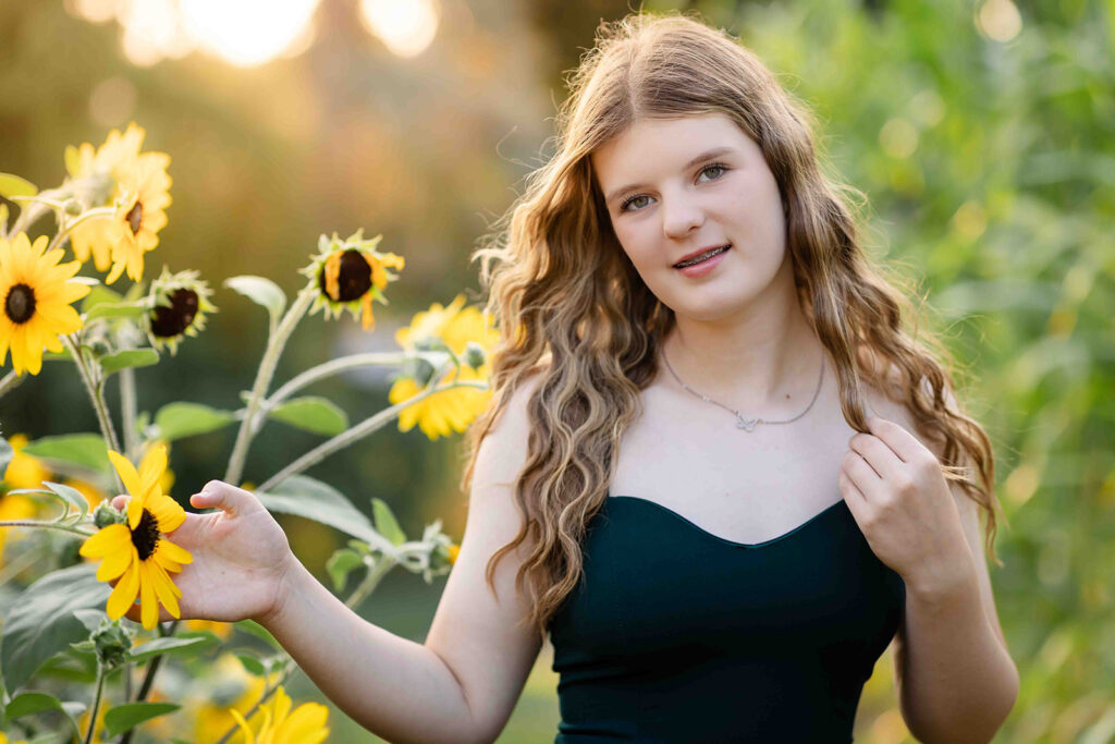 Teen girl in green dress at Rockway Gardens in Kitchener-Waterloo holding sunflower and touching hair while looking at the camera during birthday photoshoot.