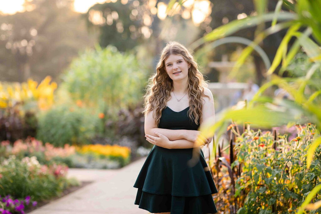Teen girl in dark green dress posing for portrait photographer in Kitchener-Waterloo with arms loosely crossed, smiling softly in a garden.