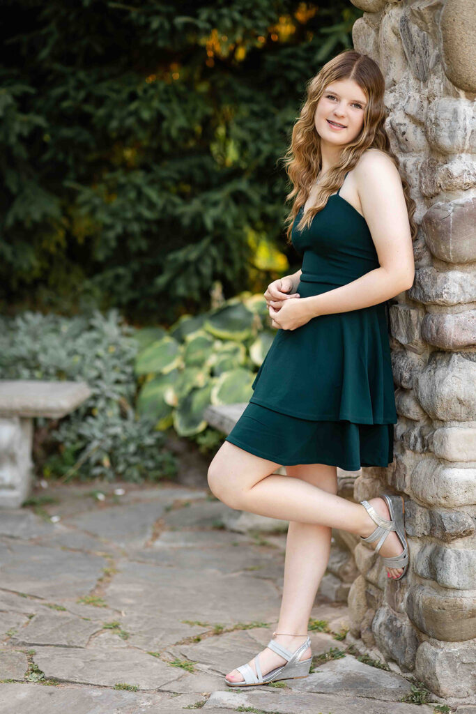 Teen girl leaning against stone wall at Rockway Gardens in Kitchener-Waterloo, smiling naturally for women’s outdoor portrait photography session.