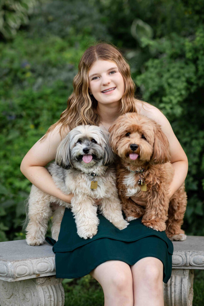Teen girl in Kitchener-Waterloo birthday photoshoot sitting on a bench holding two small fluffy dogs, both looking at the camera.