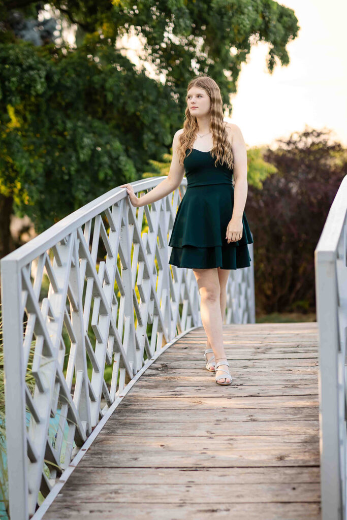 Teen girl at Rockway Gardens in Kitchener-Waterloo wearing a dark green dress, standing on a bridge with hands resting, looking poised and confident for portrait photographer session.