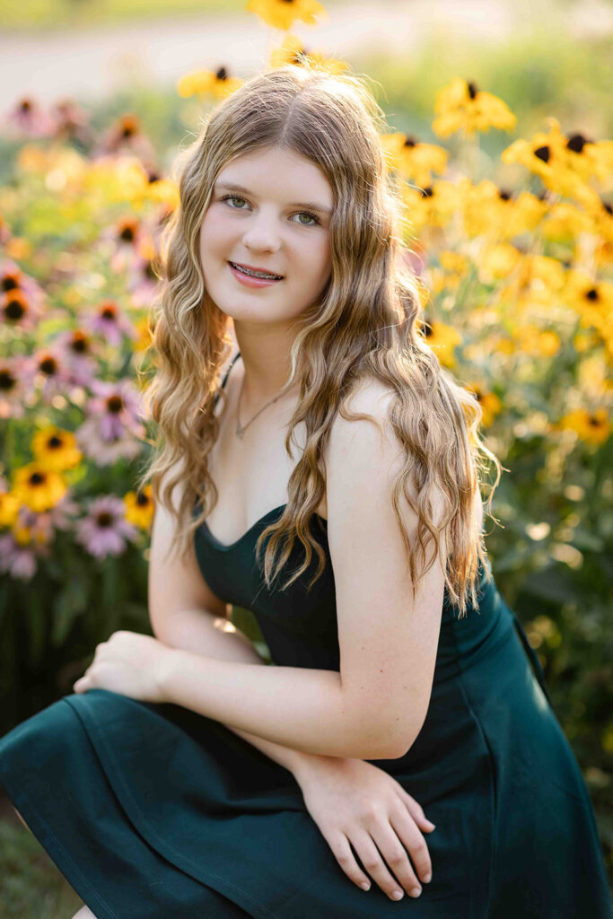 Portrait of teen girl in Kitchener-Waterloo kneeling in front of yellow flowers, smiling with soft sunlight behind her during outdoor photo session.