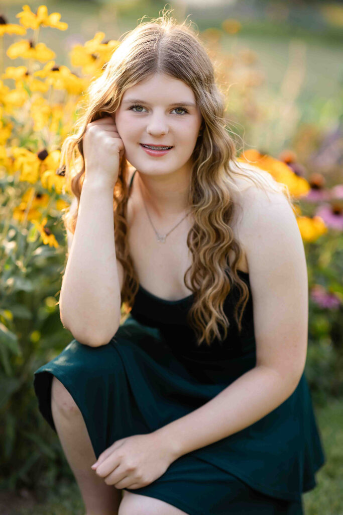 Portrait of teen girl in Kitchener-Waterloo kneeling in front of yellow flowers, smiling with soft sunlight behind her during outdoor photo session.