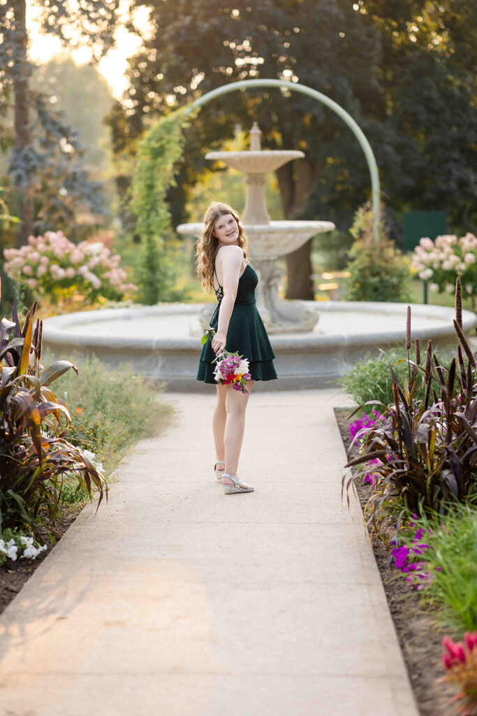 Teen girl in green dress holding a flower bouquet, looking over her shoulder with a soft smile at an outdoor portrait location in Kitchener-Waterloo.