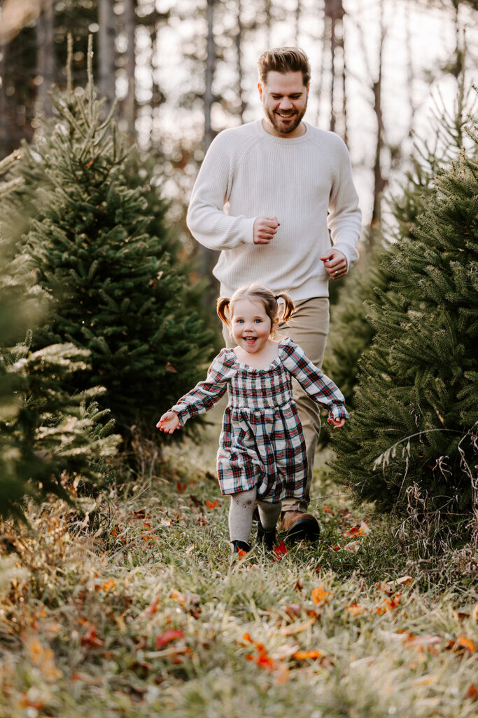 A photo of Dad chasing his daughter during holiday mini photo session at Chickadee Tree Farm featuring warm family moments among the Christmas trees — captured by Portraits by Kendra, Kitchener-Waterloo family photographer.