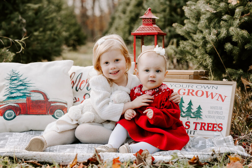 Christmas photo of two little sisters sitting on a blanket taken by Portraits by Kendra Photography at a tree farm in Kitchener-Waterloo