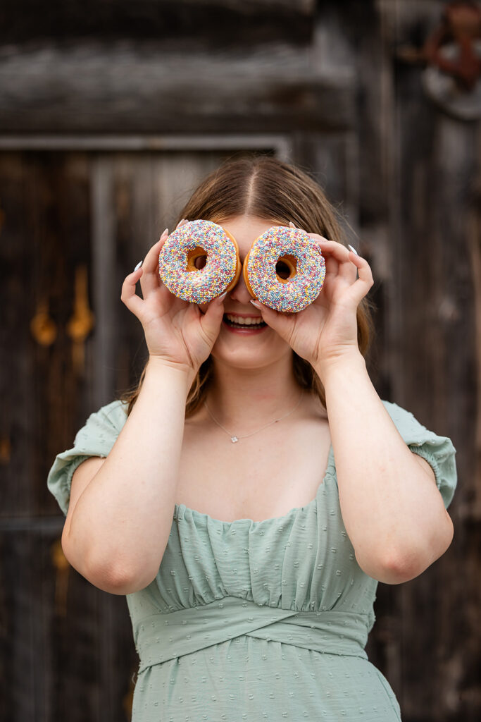 Birthday portrait of teen girl holding two donuts in front of her eyes while standing in front of barn doors. 