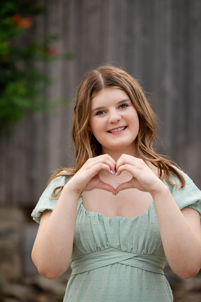 Birthday portrait of teen girl making a heart sign with her fingers, standing in front of barn doors. 